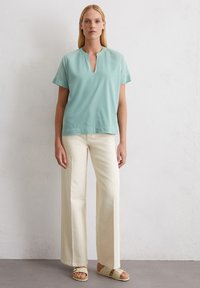 Top azzurro chiaro a maniche corte con scollatura a chiave, abbinato a pantaloni beige a gamba larga e sandali beige su uno sfondo neutro.