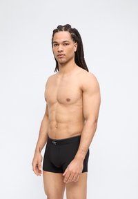 Άνδρας χωρίς πουκάμισο με μακριά πλεγμένα μαλλιά, φορώντας μαύρα εσώρουχα Calvin Klein, στέκεται μπροστά σε απλό ανοιχτό φόντο, κοιτώντας μπροστά.