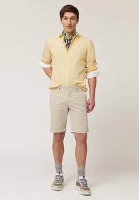 Camisa de manga larga color amarillo claro, pantalones cortos beige, calcetas grises y zapatillas casuales. Los accesorios incluyen una bufanda estampada atada al cuello.