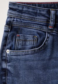 Jeans de mezclilla con un lavado azul oscuro, que cuentan con costuras amarillas en contraste, un pequeño bolsillo frontal con un acento rojo y un detalle de botón metálico.