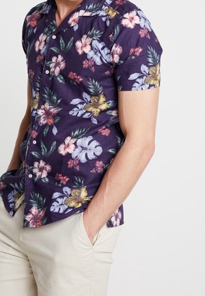 Mann trägt ein kurzärmeliges violettes Hemd mit mehrfarbigem tropischem Blumenmuster und hellbeige Hose, Hände in den Taschen.