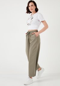 Afgeknipte witte t-shirt met bloemendesign; losse olijfgroene broek met elastische tailleband en zakken; witte sneakers.