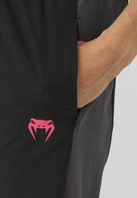 Svart stoff treningsbukser med en sidelomme som har en rosa edderkopp-logo og en glidelåsdetalj på venstre side.
