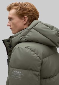 Chaqueta acolchada verde con capucha desmontable, con textura acolchada, herrajes negros y un parche de logo en el lateral. Acabado de tela suave.