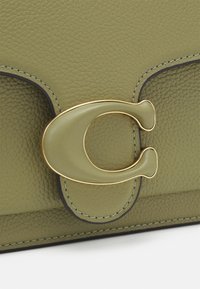 Zbliżenie na torebkę wykonaną z oliwkowej fakturowanej skóry, z dopasowanym zielonym zapięciem z złotymi akcentami w kształcie litery "C" na przedniej klapie.