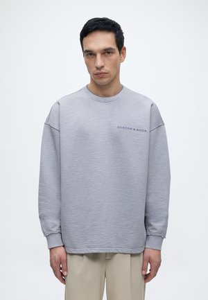 Homme portant un sweat-shirt gris clair Scotch & Soda et un pantalon beige, debout devant un fond blanc uni, face à l'avant.