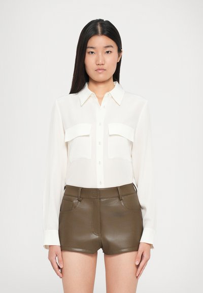 Fehér, hosszú ujjú ing gombos elülső résszel és két mellzsebbel, sötét olívazöld műbőrnadrággal párosítva, varrásrészletekkel.