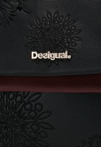 Musta teksturoidusta nahasta valmistettu laukku, jossa on kukkabossaus ja marooni yksityiskohta ylhäällä. Varustettu hopeisella "Desigual"-logolla.