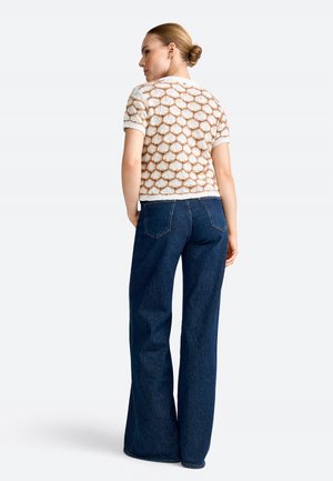 Sieviete ar matiem sasietiem kuplā sasukā valkā īsroku teksturētu džemperi ar sešstūra rakstu un tumšas platu kāju džinsas, skatās prom no kameras.