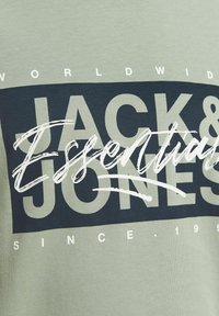 Camiseta de algodón verde con un gran gráfico azul oscuro y texto blanco, que incluye "JACK & JONES" y "Essentials", además de pequeños detalles debajo.