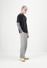 Joven con cabello rubio claro que lleva camisetas de rayas y cuadros en capas, pantalones grises, zapatillas blancas y gafas de sol futuristas, de pie de lado.