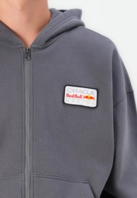 Grey zip-up hoodie with a soft texture, featuring an embroidered Oracle Red Bull Racing patch on the chest.