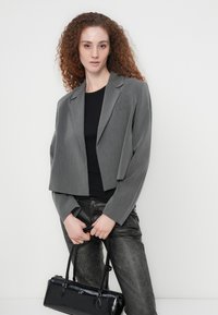 Γκρι κοντό σακάκι με αιχμηλούς γιακάδες, συνδυασμένο με μαύρο τοπ. Το μοντέλο κρατάει μία μαύρη τσάντα από πατέντ, φορώντας γκρι υφασμάτινο παντελόνι.