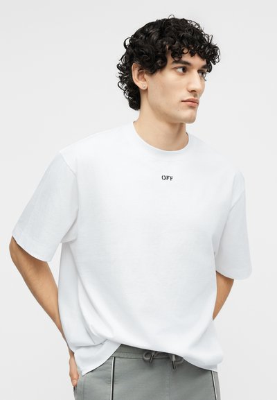 Νέος με σγουρά μαλλιά, φοράει λευκό, φαρδύ T-shirt με το κείμενο "OFF" και γκρι παντελόνι με κορδόνι, κοιτάζοντας προς τα δεξιά.