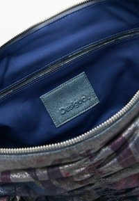 Taška so zipsom s tmavomodrou textilnou vnútornou časťou, ktorá má denimovú značku. Vonkajšok má štruktúrovaný fialovo-sivý vzorovaný povrch.