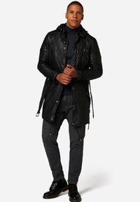 Schwarze Leder-Parka mit Kapuze, ausgestattet mit Reißverschluss- und Druckknopftaschen, strukturierten Ärmeln und einem Kordelzug in der Taille. Getragen mit dunklen Jeans und Stiefeln.