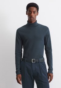 Donkergroene coltrui van gladde stof, gecombineerd met gestructureerde marineblauwe corduroybroek en een zwarte riem met een zilveren gesp.