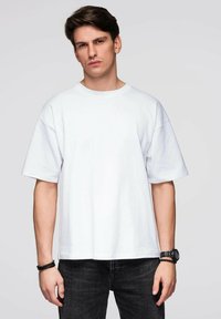 Biała, oversizeowa bawełniana koszulka z okrągłym dekoltem i krótkimi rękawami, o luźnym kroju i gładkiej fakturze. Noszona z ciemnymi dżinsami.