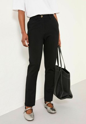 Schwarze maßgeschneiderte Hose mit einem Knopfverschluss, glattes Material, schmaler Schnitt. Kombiniert mit silbernen Ballerinas und einer schwarzen Ledertasche mit weißer Naht.