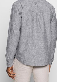 Harmaa pellavapaita, jossa pitkät hihat, napilliset kalvosimet, takahalkio ja teksturoitu viimeistely. Yhdistetty vaaleisiin housuihin.