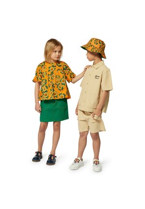 Deux enfants portent des vêtements colorés : l'un en chemise à fleurs jaune et jupe verte, l'autre en chemise beige, short et chaussures décorées.