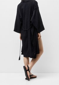Túnica de estilo kimono negra con textura, de mangas largas, ceñida en la cintura con un lazo y con una abertura lateral. Combinada con sandalias planas negras.