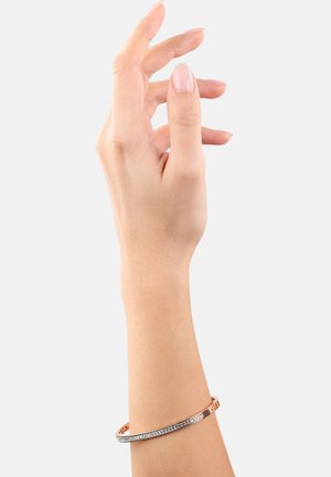 Bracciale in metallo rosato con una fila di cristalli trasparenti su un lato, esposto su una mano con dita affusolate.