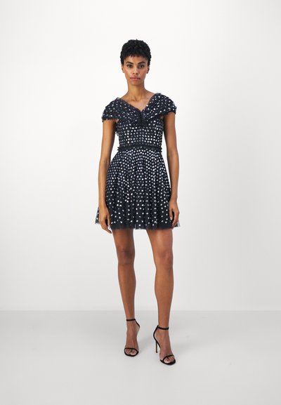 Rochie neagră cu model cu buline, umeri bufanți, corset ajustat, fustă evazată și textură din tul transparent. Asortată cu tocuri negre.