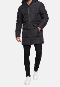 Parka noire rembourrée avec fermeture éclair, deux poches poitrine et une capuche. Portée avec un pantalon noir et des baskets blanches.