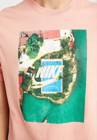 Camiseta de algodón rosa claro con un estampado gráfico aéreo de una cancha de baloncesto, con el logotipo de "NIKE" y un fondo de agua turquesa.