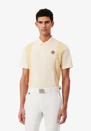 Polo color crema con pannelli laterali giallo chiaro. Presenta un piccolo logo a forma di diamante sul petto. Abbinata a pantaloni bianchi e cintura.