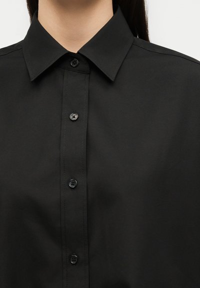 Κοντινό πλάνο ενός ατόμου που φοράει μαύρο πουκάμισο με κουμπιά και μυτερό γιακά, εστιασμένο στην περιοχή του πάνω μέρους του στήθους και του λαιμού.