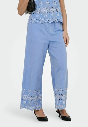 Nő világoskék, bő szárú nadrágban és hozzá illő felsőben, fehér virágos hímzéssel, fekete kézitáskát tart, fekete magassarkú cipőt visel.