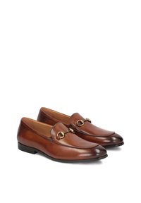 Mocasines de cuero marrón con un diseño elegante, que presentan un acabado en tono dorado en la parte delantera y una punta redondeada.