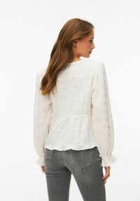 Femme aux cheveux longs bruns portant un chemisier blanc texturé à manches longues et un jean gris, de profil avec le dos partiellement visible.