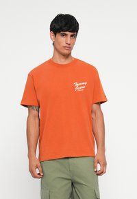 Orange cotton T-shirt with a round neckline and short sleeves. Features white “Tommy Jeans” logo on the left side and casual fit.