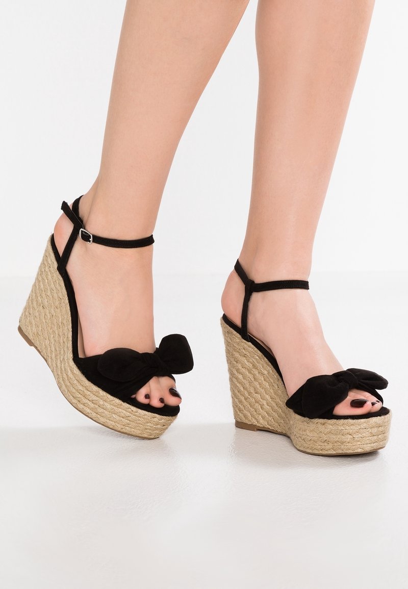Pieds chaussés de sandales compensées noires à brides autour de la cheville, avec plateforme en paille tressée et grands nœuds en tissu sur les orteils, sur fond blanc uni.