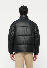 Svart dunjakke med skinnende overflata, oversized design, horisontal quilting og høy krage. Elastiske mansjetter og nederkant for en sikker passform.