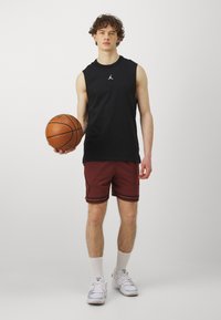 Camiseta sin mangas negra con un logo, pantalones cortos burdeos con rayas negras, sosteniendo un balón de baloncesto y usando zapatillas y calcetines deportivos blancos.