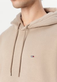 Haut à capuche beige en tissu doux avec un cordon de serrage. Présente un petit logo brodé sur la poitrine. Texture lisse et coupe régulière.