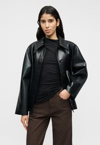 Μαύρο, γυαλιστερό μπουφάν από συνθετικό δέρμα με φαρδιά γιακά, κλείσιμο με κουμπιά και άνετη εφαρμογή, συνδυασμένο με σκούρο τοπ και καφέ παντελόνια.