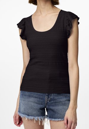 Woman wearing a fitted black textured short sleeve top with ruffled sleeves and frayed blue denim shorts.