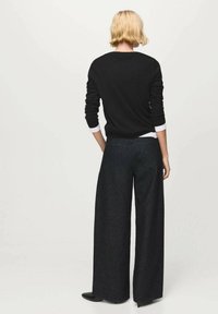 Schwarzer Pullover mit langen Ärmeln, kombiniert mit einem weißen Hemd, dazu eine dunkle, weit geschnittene Jeanshose, Ansicht von hinten.