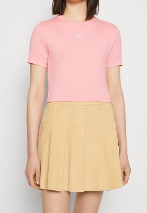 Personne portant un haut rose ajusté à manches courtes avec un petit logo blanc et une jupe plissée beige taille haute.