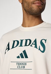 Sweatshirt crème avec un texte brodé en vert foncé : "ADIDAS" avec "TENNIS CLUB" en dessous et un logo Adidas. Encolure arrondie.