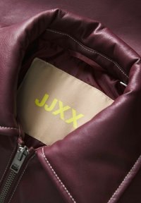 Kolaż burgundowej skórzanej kurtki z beżową podszewką i dużą żółtą etykietą z napisem "JJXX". Posiada srebrny zamek błyskawiczny oraz białe detale szewne.