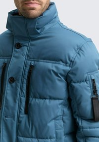Padded Jacke in Teal mit hohem Kragen. Verfügt über schwarze Reißverschlusstaschen und Knöpfe. Glattes, strukturiertes Material mit genähten Details.