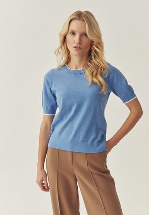 Mėlynas megztinis trumpomis rankovėmis su apvalia iškirpte ir kontrastine balta apdaila, derinamas su rudos spalvos aukšto liemens kelnėmis.