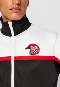Black, white, and red jacket featuring a high collar, zip closure, and a patch with a helmet design, set against a smooth fabric texture.