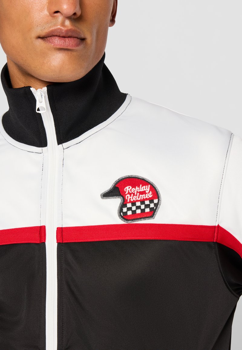 Schwarze, weiße und rote Jacke mit hohem Kragen, Reißverschluss und einem Patch mit einem Helmdesign, vor einer glatten Stoffstruktur.